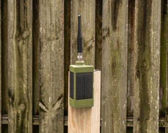May include: A rectangular olive green electronic device featuring a black solar panel on its front face and a vertical black antenna extending from the top center. The unit is mounted on a light-colored wooden post with a smooth, finished surface.