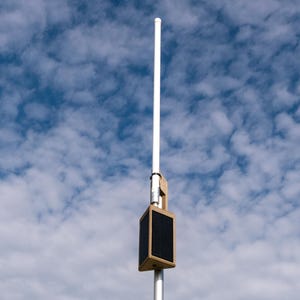 May include: A tall, slender white antenna pole mounted with a rectangular, tan-colored solar-powered device. The device features a dark, flat solar panel surface on its side, securely attached to the vertical pole against a blue sky.