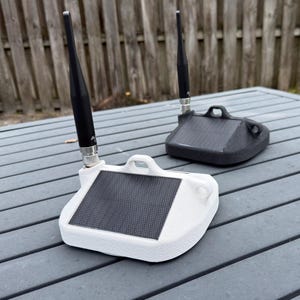 May include: Two wireless sensor devices, one white and one black, each featuring a textured base, a central rectangular solar panel, a small integrated handle, and a tall black antenna attached via a metallic connector to the top of the unit.