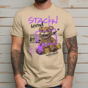 Stackin Bread Teddy Graphic T-Shirt in Sand featuring a bold teddy bear design with vibrant purple accents. This trendy streetwear shirt is perfect for men and women who love urban fashion. Made with soft fabric for all-day comfort and bold style.