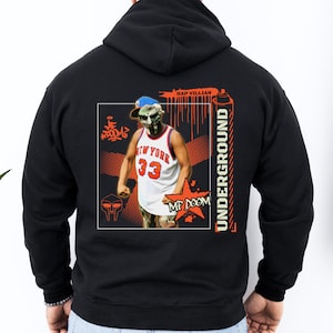 May include: Black hooded sweatshirt with a graphic print of a cartoon character wearing a basketball jersey with the text "New York" and "33" on the front. The character is holding a spray can and the text "MF DOOM" is in a red star. The text "RAP VILLIAN UNDERGROUND" is on the right side of the graphic.