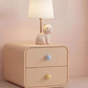 May include: A beige nightstand with two drawers, each with a rounded blue and yellow knob. A white cat figurine sits atop the nightstand, with a small lamp with a white shade and gold stem behind it.