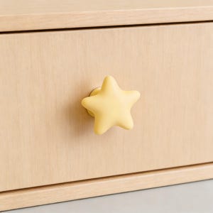 Op de afbeelding: Een close-up van een lichtgekleurde houten lade met een gele stervormige knop. De knop is gecentreerd op de voorkant van de lade en zorgt voor een speels en decoratief element. De houtnerf is zichtbaar.