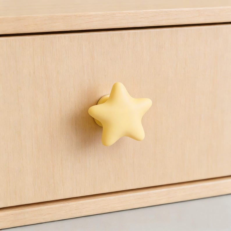 Op de afbeelding: Een close-up van een lichtgekleurde houten lade met een gele stervormige knop. De knop is gecentreerd op de voorkant van de lade en zorgt voor een speels en decoratief element. De houtnerf is zichtbaar.