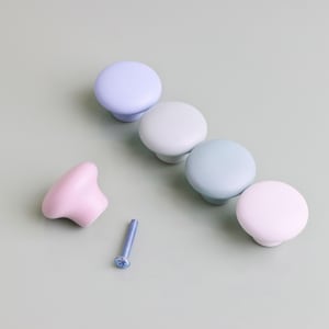 Op de afbeelding: Een verzameling pastelkleurige kastknoppen in tinten lavendel, mintgroen en roze. E&eacute;n roze knop staat los van de andere, met een blauwe schroef in de buurt. De knoppen zijn rond met een platte bovenkant en een korte, afgeronde basis. De achtergrond is een gedempt groen.