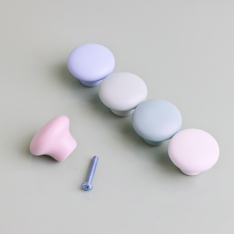 Op de afbeelding: Een verzameling pastelkleurige kastknoppen in tinten lavendel, mintgroen en roze. E&eacute;n roze knop staat los van de andere, met een blauwe schroef in de buurt. De knoppen zijn rond met een platte bovenkant en een korte, afgeronde basis. De achtergrond is een gedempt groen.