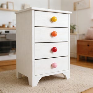 May include: A white wooden bedside cabinet with four drawers, each with a brightly coloured round knob. The knobs are yellow, orange, red, and pink. The cabinet has a distressed finish and is on a light-coloured rug.