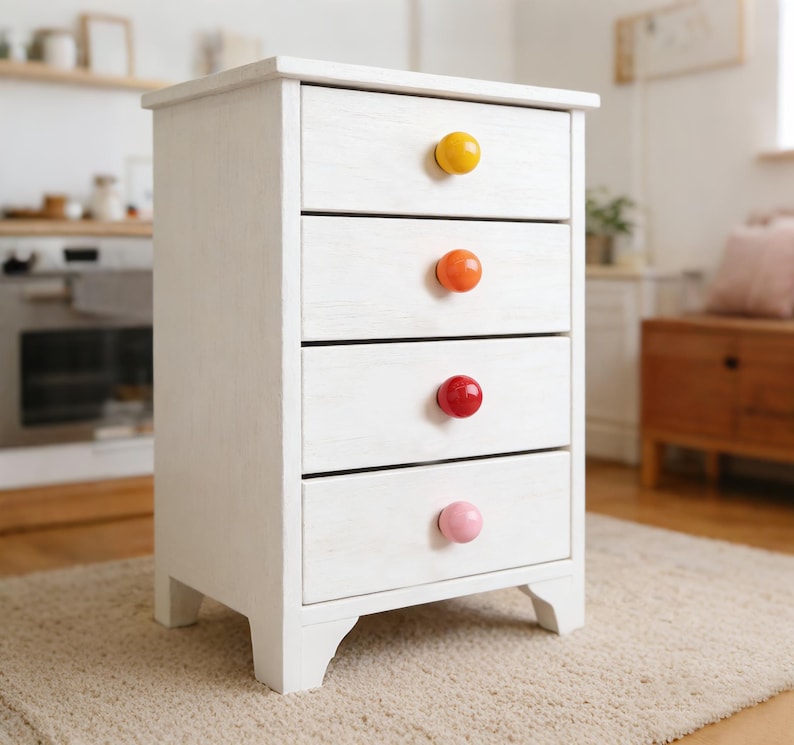 May include: A white wooden bedside cabinet with four drawers, each with a brightly coloured round knob. The knobs are yellow, orange, red, and pink. The cabinet has a distressed finish and is on a light-coloured rug.