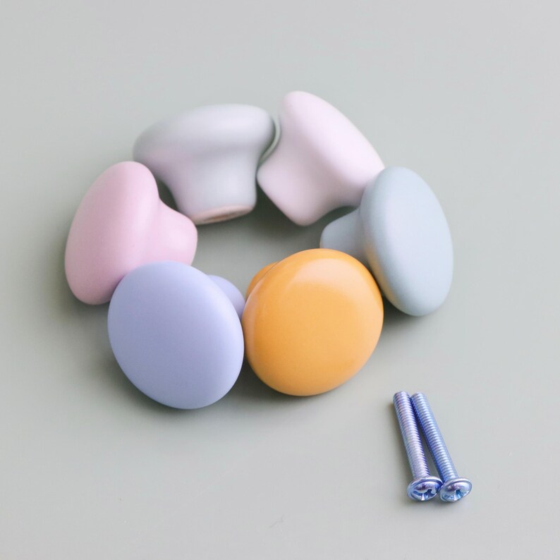 Op de afbeelding: Een verzameling pastelkleurige kastknoppen in roze, blauwe, gele en groene tinten, gerangschikt in een cirkel. Twee zilveren schroeven zijn naast de knoppen geplaatst. De knoppen hebben een glad, afgerond ontwerp.