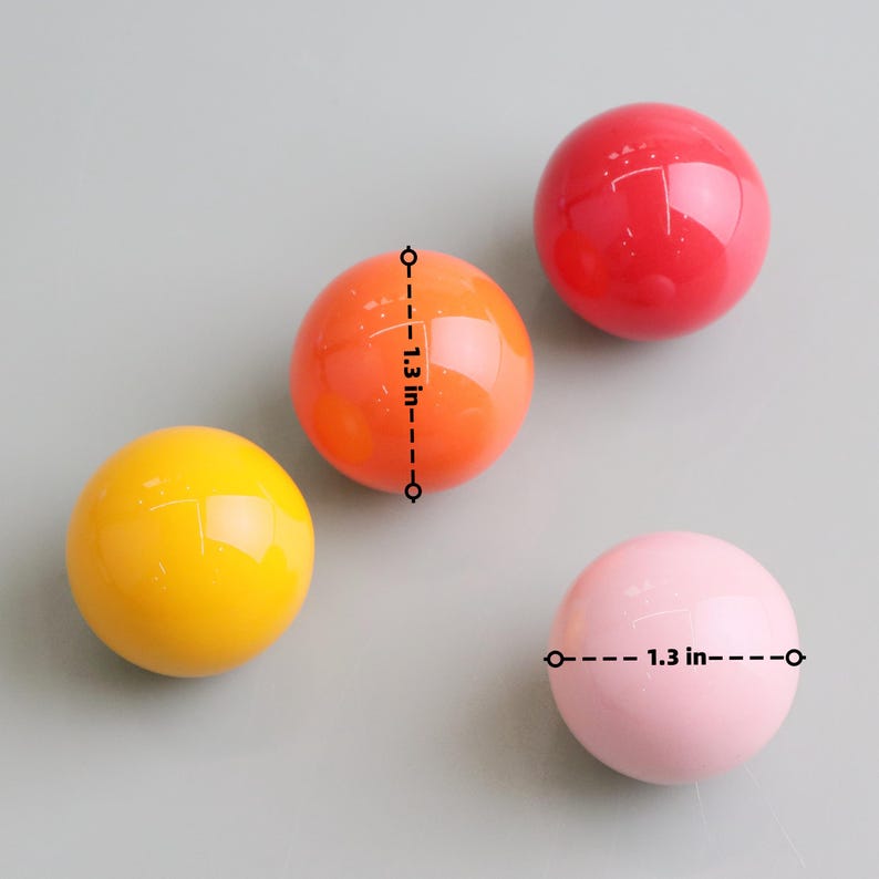 May include: Four glossy, spherical objects in yellow, orange, red, and pink. Each ball is approximately 3.3 cm in diameter. The objects are arranged on a light grey surface, showcasing their vibrant colours and smooth texture.