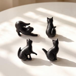 May include: A set of four small, black, cast metal cat-shaped knobs. Each knob features a different feline pose, including one cat stretching, one sitting upright, and two in various seated positions, all with a textured, matte black finish.