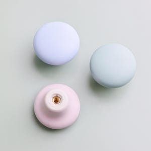 Op de afbeelding: Drie pastelkleurige kastknoppen. De knoppen zijn rond met een matte afwerking. E&eacute;n is lichtpaars, &eacute;&eacute;n is mintgroen en de derde is roze. De knoppen zijn waarschijnlijk gemaakt van keramiek of porselein en zijn geschikt voor meubels.