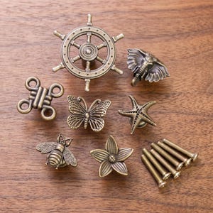 Puede incluir: Una colección de pomos para armarios de metal en tono latón antiguo con diversas formas, incluyendo un timón de barco, cabeza de elefante, mariposa, estrella de mar, abeja, flor y un nudo decorativo, junto con seis tornillos metálicos a juego.