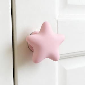Op de afbeelding: Een lichtroze stervormige deurknop. De knop is bevestigd op een witte deur. De ster heeft afgeronde randen en een gladde, matte afwerking. De deurknop is een decoratief element.