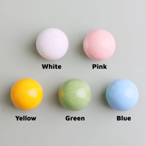 May include: Five glossy, round knobs in white, pink, yellow, green, and blue. Each knob is labelled with its colour name in a bold, black font. The knobs are arranged on a light grey background.
