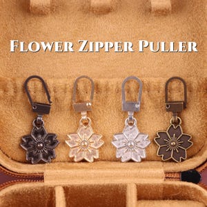 Puede incluir: Cuatro tiradores de cremallera con forma de flor en diferentes acabados metálicos: bronce oscuro, dorado, plateado y bronce antiguo. Cada tirador presenta un diseño floral y un pequeño cierre metálico. Se muestra el texto "FLOWER ZIPPER PULLER".