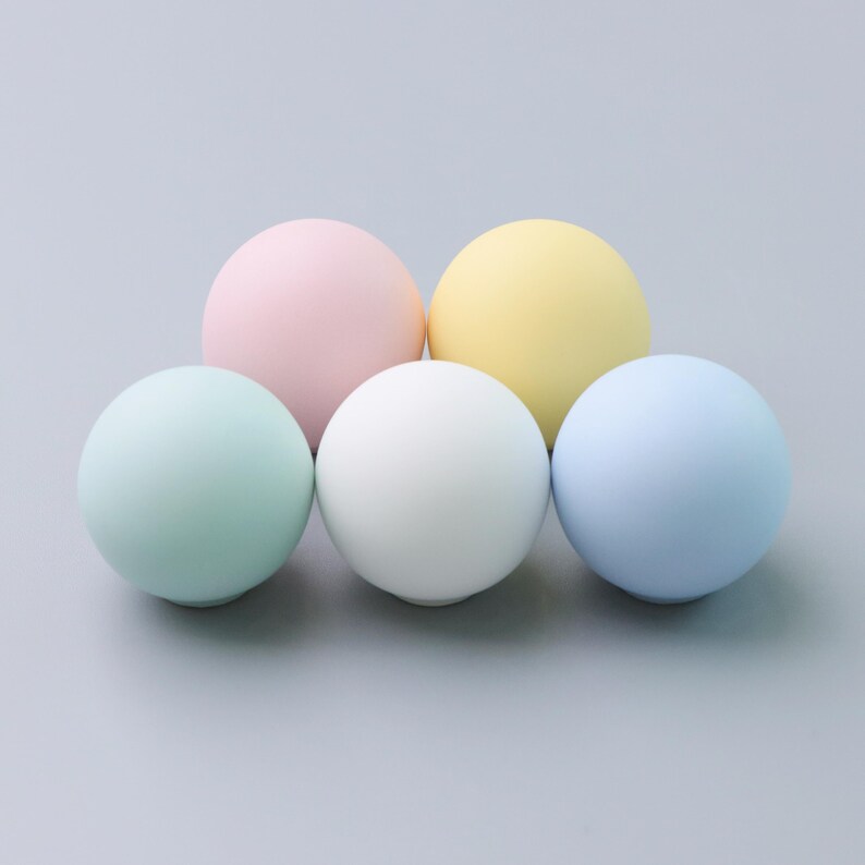 May include: Five pastel-colored spheres arranged on a gray surface. The spheres are in shades of pink, yellow, white, blue, and green. The smooth, round objects are evenly spaced and appear to be made of a matte material.