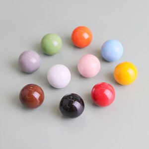 May include: Eleven glossy, spherical knobs in a variety of colours: brown, black, red, pink, orange, yellow, green, blue, white, and lavender. The decorative knobs are arranged on a light grey surface.