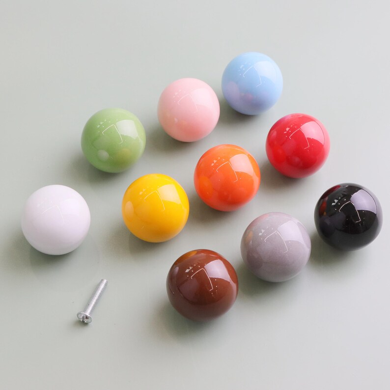 May include: A selection of spherical cabinet knobs in a range of colours: white, green, pink, yellow, orange, red, grey, brown, blue, and black. A silver screw is also visible. These decorative knobs are likely for furniture.