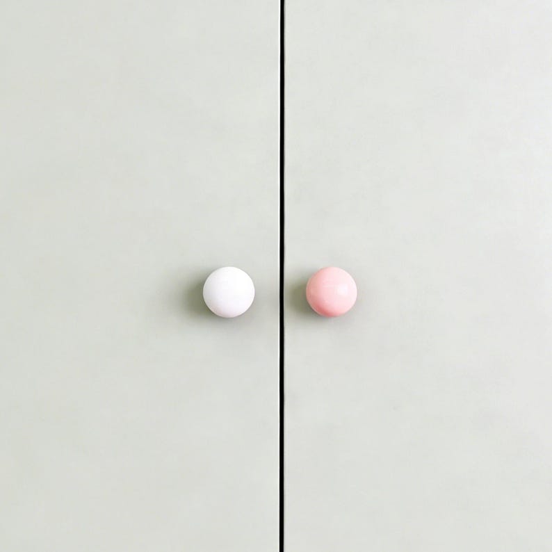 May include: A close-up of a light green cabinet door with two round knobs. One knob is white, and the other is pink. The knobs are centered on the door panels.