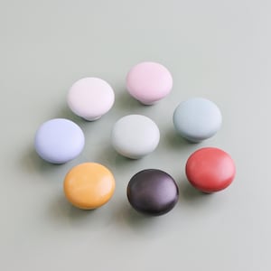 Op de afbeelding: Een verzameling van acht kleurrijke keramische kastknoppen in verschillende pastel- en felle kleuren, waaronder wit, roze, blauw, groen, geel, zwart en rood. De knoppen zijn rond en hebben een gladde, glanzende afwerking. Diameter ongeveer 3 cm.