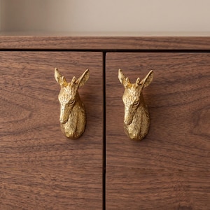 May include: Two gold-colored deer head cabinet pulls mounted on a dark brown wooden cabinet. The deer heads are detailed with antlers and facial features. The cabinet has a natural wood grain.