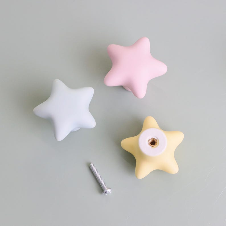 Op de afbeelding: Drie stervormige kastknoppen in pastelkleuren: lichtblauw, roze en geel. Er is ook een zilveren schroef aanwezig. Deze decoratieve knoppen zijn ideaal om een speelse toets aan meubels toe te voegen.