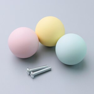 May include: Three pastel-colored cabinet knobs in pink, yellow, and blue, with two silver screws. The knobs are spherical and smooth, ideal for adding a pop of color to furniture. The hardware is set against a light gray background.
