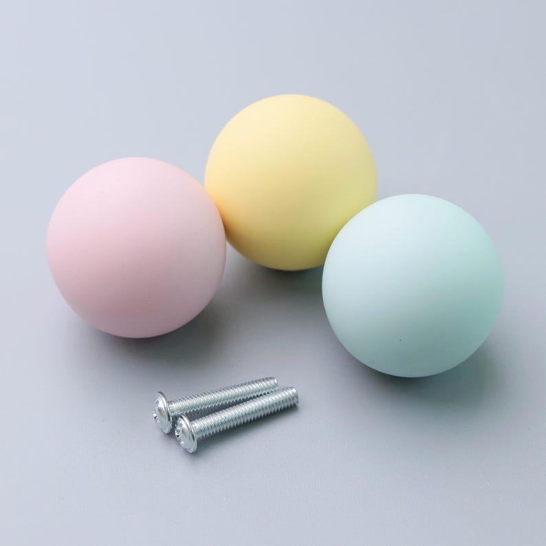 May include: Three pastel-colored cabinet knobs in pink, yellow, and blue, with two silver screws. The knobs are spherical and smooth, ideal for adding a pop of color to furniture. The hardware is set against a light gray background.