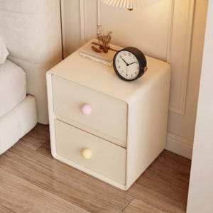 May include: A cream-colored nightstand with two drawers, each with a round knob. The top of the nightstand holds a black analog clock, a small glass, and a decorative item. The nightstand is next to a bed.