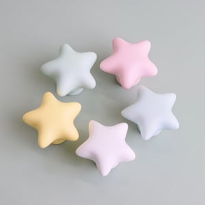 Op de afbeelding: Zes stervormige kastknoppen in pastelkleuren: geel, roze, lichtblauw en lichtgroen. De knoppen zijn in een cirkelvormig patroon op een lichtgroen oppervlak gerangschikt. Deze decoratieve knoppen zijn ideaal voor kindermeubels.