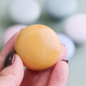 Op de afbeelding: Een hand houdt een glad, rond, mosterdgeel object vast. Het object is scherp, met een glanzend oppervlak. Zachte pastelkleurige bollen zijn op de achtergrond. De hand heeft donkere nagellak.
