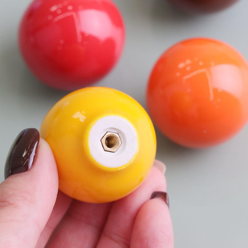 May include: Close-up of several colourful, round cabinet knobs. One yellow knob is in focus, held by a hand, with a white base and a brass screw. Other knobs are red, orange, and brown, all with a glossy finish. The knobs are for furniture.
