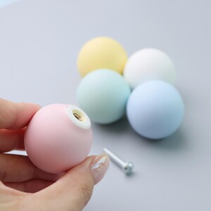 May include: A pastel pink spherical cabinet knob with a metal insert is held in a hand. Four other spherical knobs in pastel yellow, white, mint green, and light blue are in the background. A silver screw is also visible.