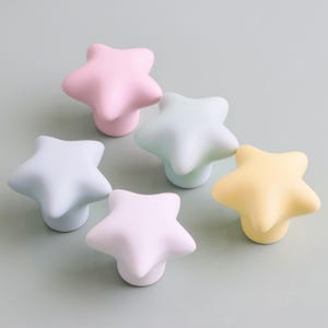 Op de afbeelding: Een verzameling van vijf stervormige kastknoppen in pastelkleuren: roze, blauw, groen en geel. Elke knop heeft een afgeronde basis en een vijfpuntig sterontwerp, perfect om een ​​gril te geven aan meubels.