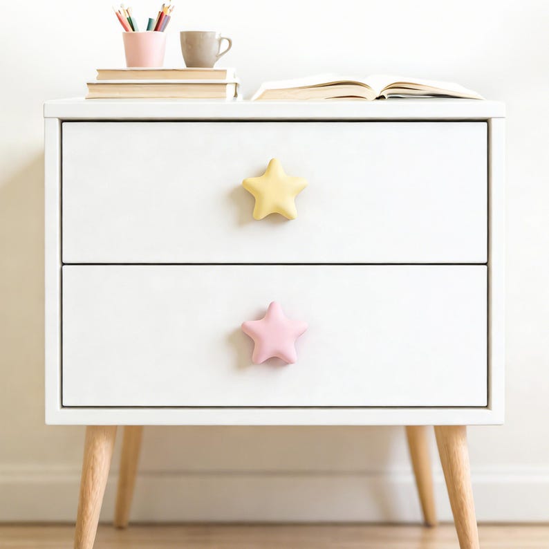 Op de afbeelding: Een witte nachtkast met twee lades, elk met een stervormige knop. De bovenste knop is geel, de onderste roze. De tafel heeft lichtgekleurde houten poten. Boeken, potloden en een mok staan bovenop.