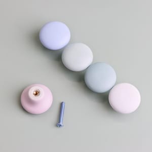 Op de afbeelding: Een verzameling pastelkleurige kastknoppen in tinten lavendel, mintgroen en roze. E&eacute;n knop is losgemaakt, met een schroef ernaast. De knoppen zijn rond en glad, perfect om een vleugje kleur aan meubels toe te voegen.