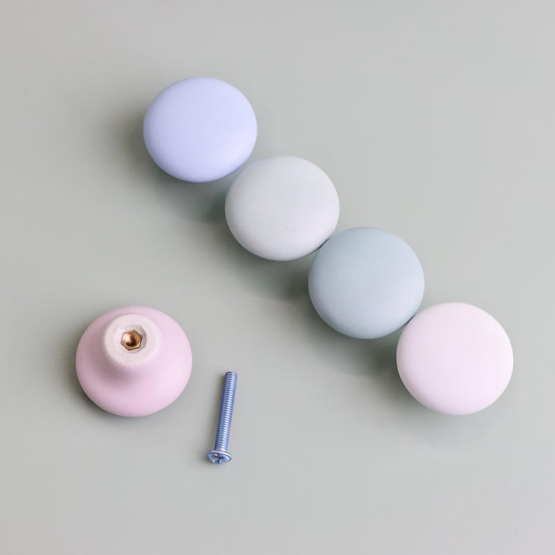 Op de afbeelding: Een verzameling pastelkleurige kastknoppen in tinten lavendel, mintgroen en roze. E&eacute;n knop is losgemaakt, met een schroef ernaast. De knoppen zijn rond en glad, perfect om een vleugje kleur aan meubels toe te voegen.