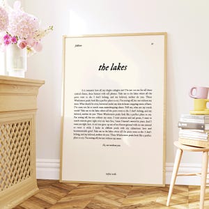 May include: A framed print featuring the text "the lakes" in a stylish font. The print includes a poem by Taylor Swift, with a cream-colored background and a wooden frame. A vase of pink flowers is visible.