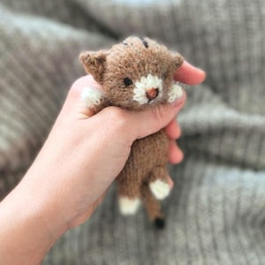 Op de afbeelding: Een klein, handgemaakt, bruin en wit gebreid kattenspeeltje. De kat heeft een witte snuit, poten en staartpunten. Het speelgoed wordt in een hand gehouden tegen een zachte, grijze achtergrond.