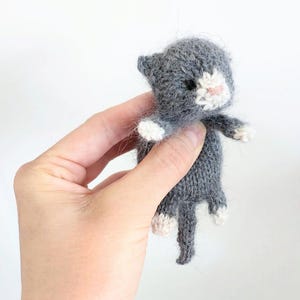 Op de afbeelding: Een handgemaakte, gebreide kattenpop in grijs en wit. De kat heeft een grijze body, kop en staart, met witte poten en een witte vlek op het gezicht. De pop wordt tegen een witte achtergrond gehouden.