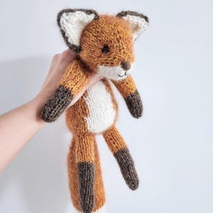 May include: A knitted fox toy in orange, brown, and white. The fox has a white chest, brown legs and tail tips, and white-tipped ears. The toy is held up against a plain white background.