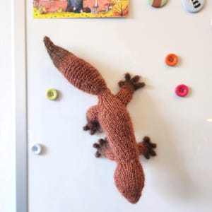 Könnte beinhalten: Eine handgestrickte braune Eidechse mit dunkelbraunen Füßen und Schwanz ist an einer weißen Oberfläche befestigt. Die Eidechse besteht aus einem weichen, strukturierten Garn. Mehrere bunte Knöpfe und ein dekoratives Bild sind ebenfalls an der Oberfläche befestigt.