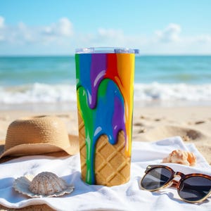 Op de afbeelding: Een hoge, geïsoleerde tumbler met een doorzichtig deksel, met een levendig ontwerp van regenboogkleurige verf die over een wafelpatroon druipt. De tumbler staat op een strand met een strohoed en een zonnebril.