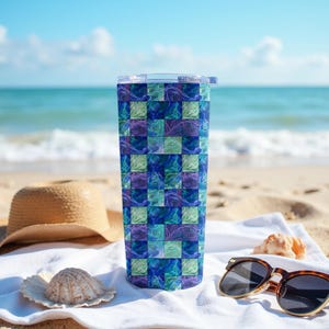 May include: A tall, insulated tumbler with a colorful, geometric pattern in shades of blue, green, and purple. The tumbler has a clear lid and is set on a white towel with sunglasses, a seashell, and a straw hat on a sandy beach.