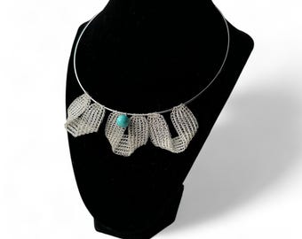 May include: A silver-coloured necklace with a rigid collar and a series of woven, petal-like dangles. A small, round, turquoise-coloured bead is centered at the bottom. The necklace is displayed on a black mannequin.