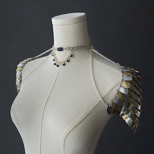 Puede incluir: Una pieza de hombro de malla de cadena plateada y dorada con un collar de cadena plateada con un colgante de piedra azul.