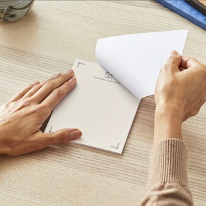 May include: A white notepad with a black border and the text "The Meaning of Life Remains" printed on the cover. The notepad is open and the top page is being turned over.