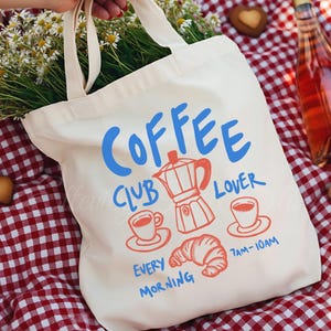 May include: A white ceramic mug with a black handle and the text "But First, Coffee" in a bold, black font. The mug is set against a blurred background, suggesting a cozy setting.