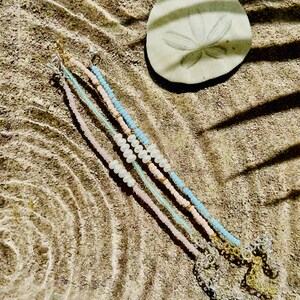 May include: Four beaded bracelets with pearl accents. The bracelets are in shades of pink, blue, and white. The bracelets are laid out on a sandy surface.
